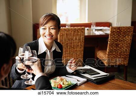 Picture - Two businesswomen sitting at table, toasting with drinks. Fotosearch