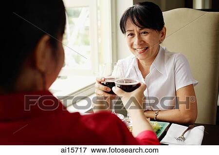 Stock Photo - Two women at restaurant, sitting face to face, toasting with wine glasses. Fotosearch