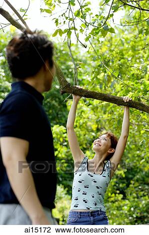 Stock Image - Woman hanging onto tree branch, man watching her. Fotosearch