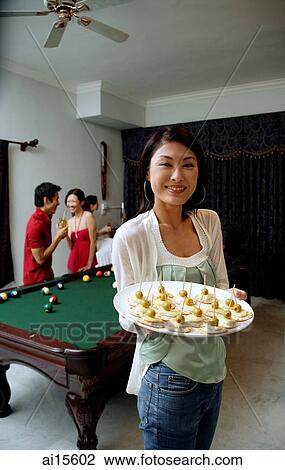 Stock Image - Woman holding tray of appetizers, smiling at camera, people in the background. Fotosearch