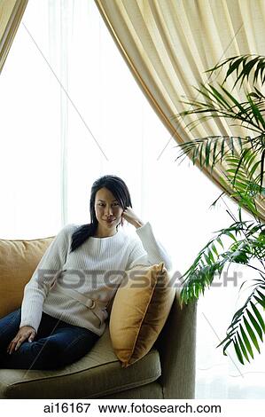 Stock Photo - Woman in living room, sitting on sofa, looking at camera. Fotosearch