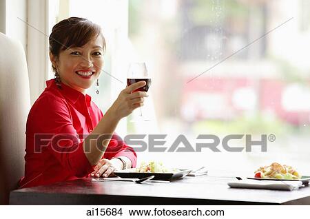 Woman sitting at table, raising drink towards camera, smiling View Large Photo Image Picture - Woman sitting at table, raising drink towards camera, smiling. Fotosearch