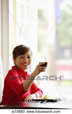 Stock Image - Woman sitting at table, raising drink towards camera. Fotosearch