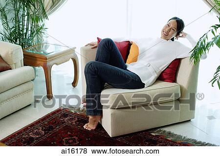 Stock Photo - Woman sitting on sofa in living room, smiling at camera. Fotosearch
