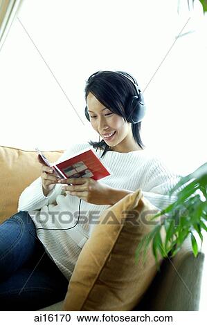 Stock Image - Woman sitting on sofa, wearing headphones, looking at CD case. Fotosearch