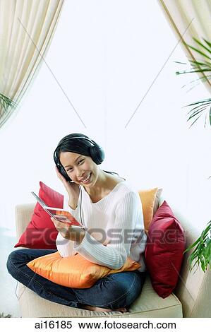 Stock Photography - Woman sitting on sofa, wearing headphones, smiling at camera, portrait. Fotosearch