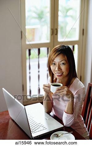 Woman using laptop, holding cup, smiling at camera View Large Photo Image Stock Photograph - Woman using laptop, holding cup, smiling at camera. Fotosearch