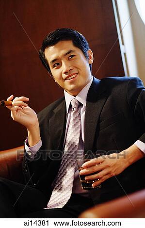 Stock Image - Young businessman holding cigar and glass of whiskey, looking away. Fotosearch