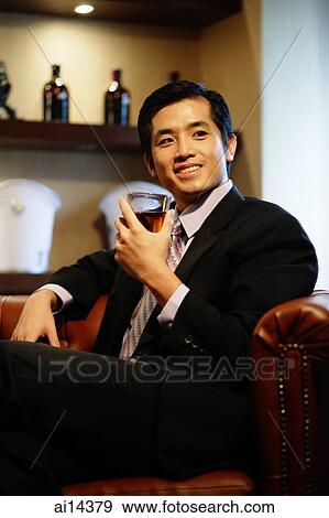 Stock Photo - Young businessman sitting on sofa, holding glass of whiskey, looking away. Fotosearch