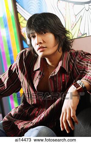 Stock Image - Young man sitting, looking at camera, portrait. Fotosearch