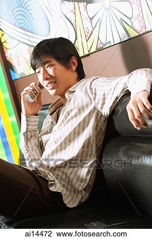 Stock Image - Young man sitting on sofa, using mobile phone. Fotosearch
