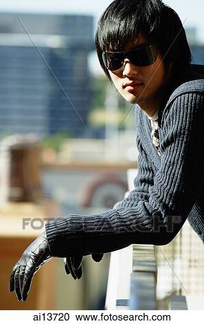 Stock Image - Young man wearing sunglasses, leaning on railing, looking at camera. Fotosearch