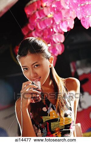 Stock Photo - Young woman holding glass to mouth, looking at camera. Fotosearch