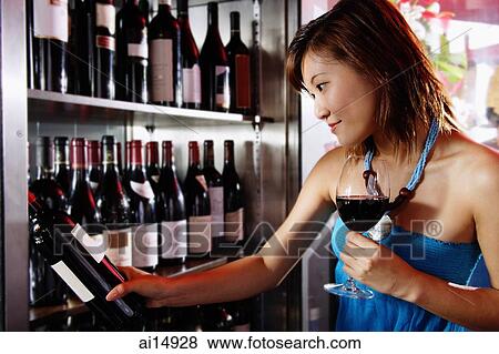 Young woman holding wine glass, looking at wine bottle on shelf View Large Photo Image Stock Photo - Young woman holding wine glass, looking at wine bottle on shelf. Fotosearch