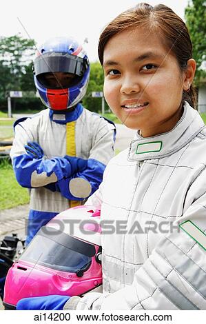 Stock Image - Young woman in sports jumpsuit, holding helmet, looking at camera, man behind her. Fotosearch