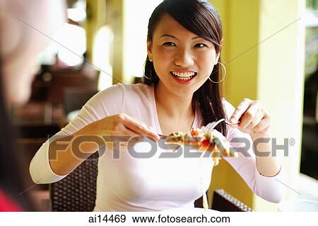 Stock Photo - Young woman lifting slice of pizza with cutlery, smiling at another person in front of her. Fotosearch