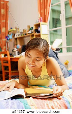 Stock Photo - Young woman, lying on bed looking at magazine. Fotosearch