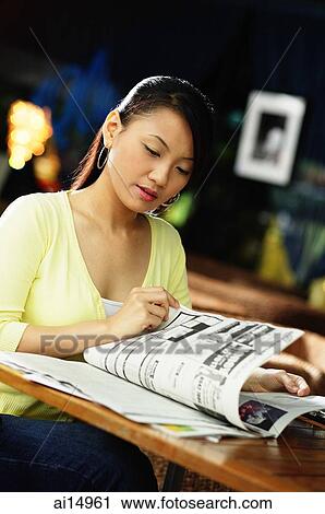 Stock Image - Young woman sitting at table, reading newspaper. Fotosearch