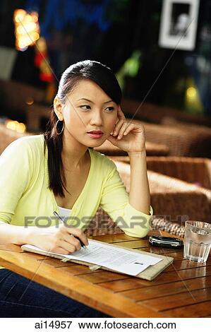 Young woman sitting at table with pen and paper, hand on face View Large Photo Image Stock Photo - Young woman sitting at table with pen and paper, hand on face. Fotosearch