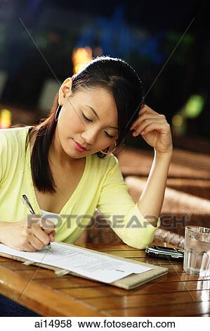 Stock Photo - Young woman sitting at table with pen and paper, writing. Fotosearch