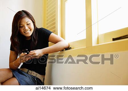Picture - Young woman sitting next to window, looking at mobile phone. Fotosearch
