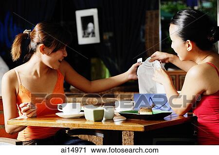 Stock Image - Young women at cafe, looking at items from shopping bag. Fotosearch