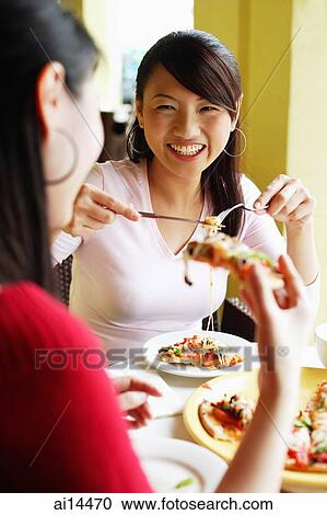 Stock Image - Young women eating pizza at cafe, over the shoulder view. Fotosearch