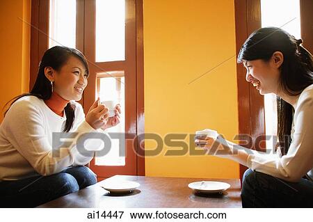 Stock Photo - Young women, holding cups of coffee, sitting face to face. Fotosearch