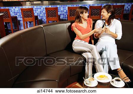 Picture - Young women sitting at cafe, one holding mobile phone. Fotosearch