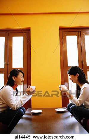 Stock Photography - Young women sitting in living room opposite each other, holding cups of coffee. Fotosearch