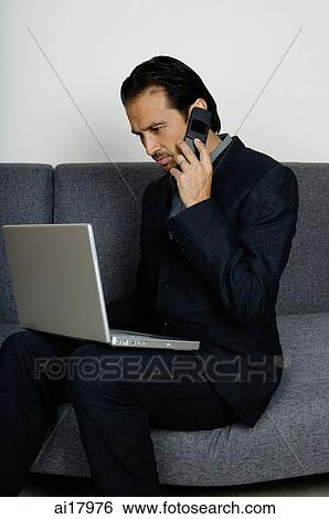 Businessman sitting on sofa, using mobile phone and looking at laptop View Large Photo Image Stock Photograph - Businessman sitting on sofa, using mobile phone and looking at laptop . Fotosearch