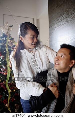 Picture - Couple in winter wear looking at each other, Christmas tree behind them . Fotosearch