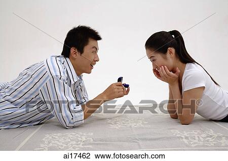 Stock Image - Couple lying on front, face to face, man holding ring box, woman with hands on chin . Fotosearch