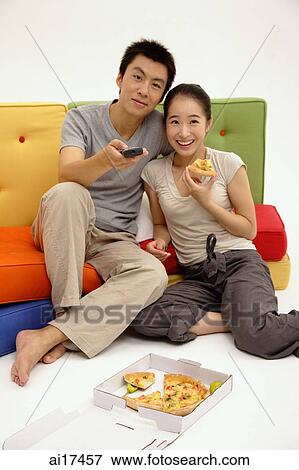 Couple sitting on sofa, woman eating pizza, man holding TV remote control View Large Photo Image Stock Photo - Couple sitting on sofa, woman eating pizza, man holding TV remote control . Fotosearch