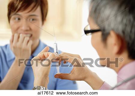 Stock Image - Doctor preparing syringe, patient cringing in background . Fotosearch