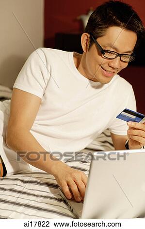 Man lying on bed, using laptop, holding credit card View Large Photo Image Stock Image - Man lying on bed, using laptop, holding credit card . Fotosearch
