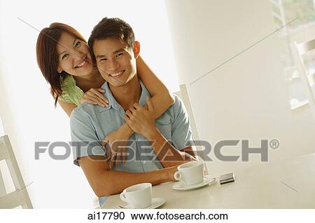 Man sitting at table, woman embracing him from behind, both smiling at camera View Large Photo Image Stock Image - Man sitting at table, woman embracing him from behind, both smiling at camera . Fotosearch