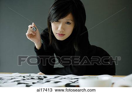 Stock Image - Woman holding paintbrush, leaning on table, looking at camera . Fotosearch
