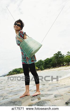 Stock Image - Woman standing on beach, carrying beach bag . Fotosearch