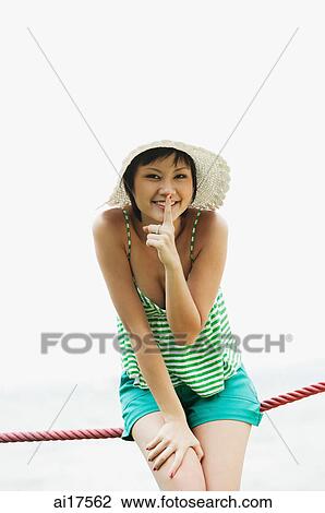 Stock Image - Woman wearing hat, smiling at camera, finger on lips . Fotosearch
