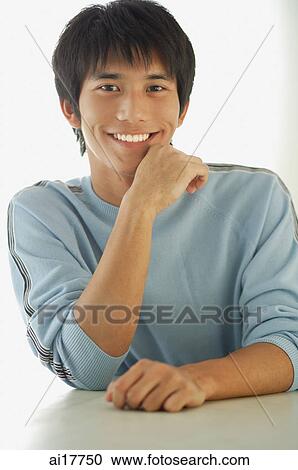Young man sitting at table, smiling at camera, hand on chin View Large Photo Image Stock Image - Young man sitting at table, smiling at camera, hand on chin . Fotosearch