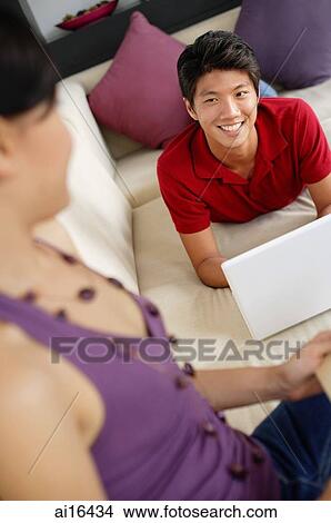 Couple in living room, man lying on sofa with laptop, smiling at woman View Large Photo Image Picture - Couple in living room, man lying on sofa with laptop, smiling at woman . Fotosearch