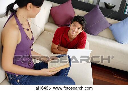 Couple in living room, man lying on sofa with laptop, woman holding book View Large Photo Image Stock Photography - Couple in living room, man lying on sofa with laptop, woman holding book . Fotosearch