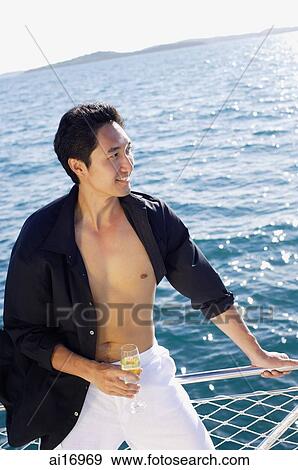 Stock Photo - Man on boat, leaning on railing, drink in hand, looking away, smiling. Fotosearch