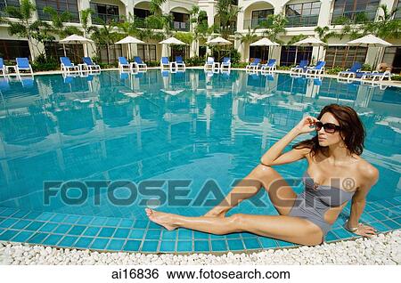 Stock Photograph - Woman in swimwear, sitting next to swimming pool, building in the background . Fotosearch