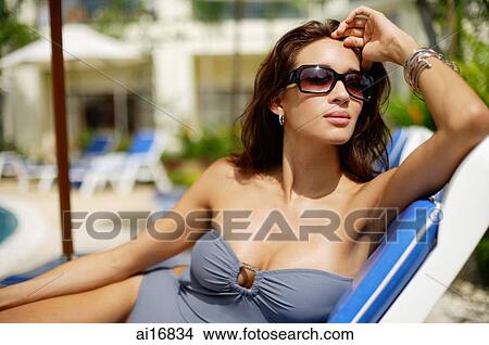 Woman in swimwear, wearing sunglasses, sitting on deck chair, hand on head View Large Photo Image Picture - Woman in swimwear, wearing sunglasses, sitting on deck chair, hand on head . Fotosearch