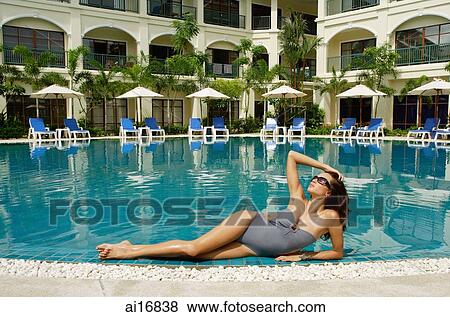 Stock Photo - Woman lying next to swimming pool, hand on head, building in the background . Fotosearch