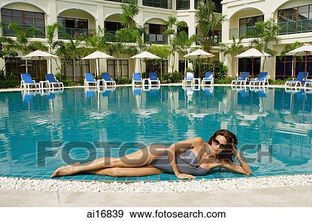 Stock Photo - Woman lying next to swimming pool, leaning on hand, building in the background . Fotosearch