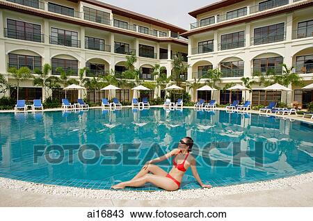 Stock Image - Woman sitting next to swimming pool, wearing bikini, hand on knee . Fotosearch