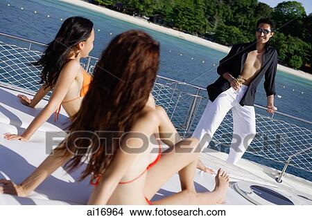 Women in bikinis sitting on boat deck, man standing, looking at them View Large Photo Image Picture - Women in bikinis sitting on boat deck, man standing, looking at them . Fotosearch